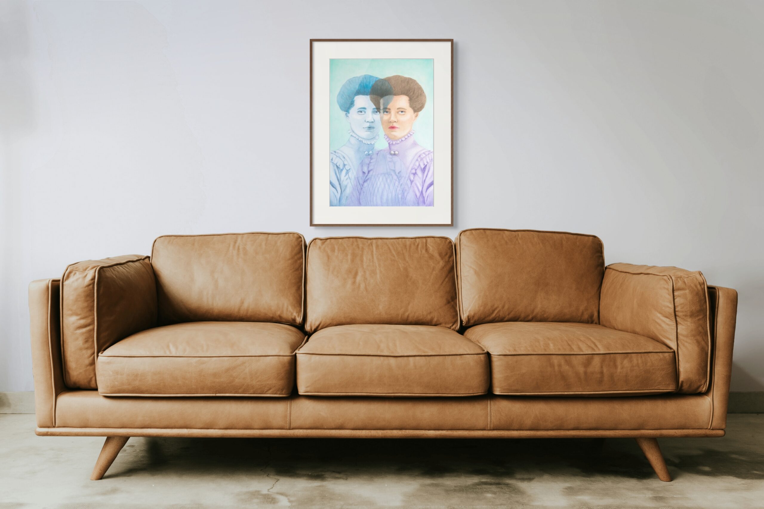 Picture of two mirrored ladies in pastel colors hanging above a brown leather sofa.